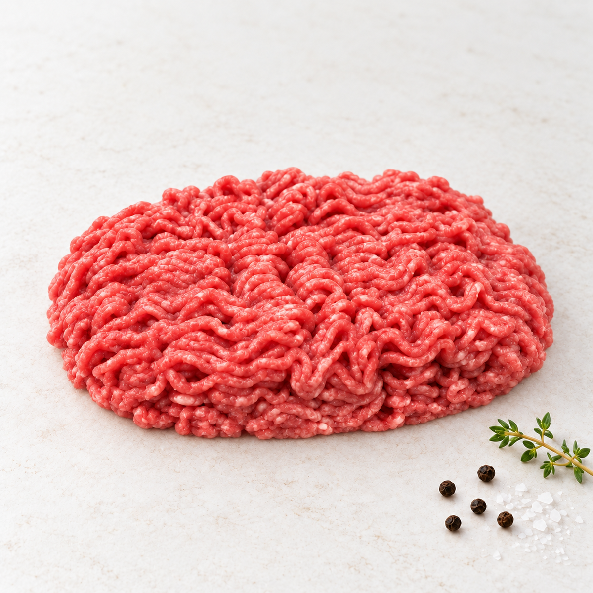 Raw lean ground beef in a rounded mound on a light stone surface with thyme, sea salt and black peppercorns.