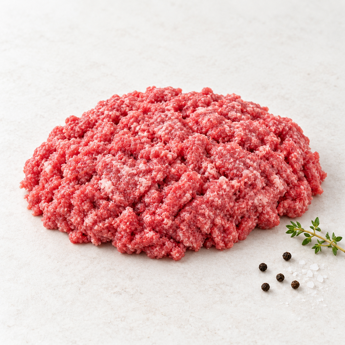 Raw Wagyu ground beef on a light stone surface with thyme, sea salt and black peppercorns.
